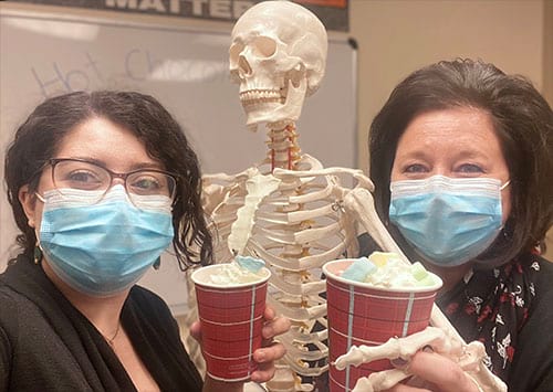Two women wearing face masks are holding cups of hot chocolate next to a skeleton.
