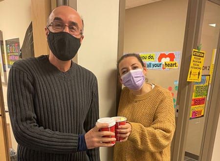 A man and a woman wearing masks are standing next to each other holding cups of coffee.