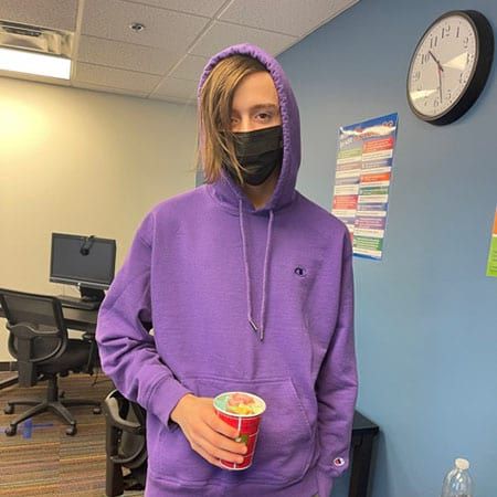 A person wearing a purple hoodie and a mask is holding a cup.