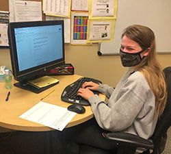 A woman wearing a mask is sitting at a desk using a computer.