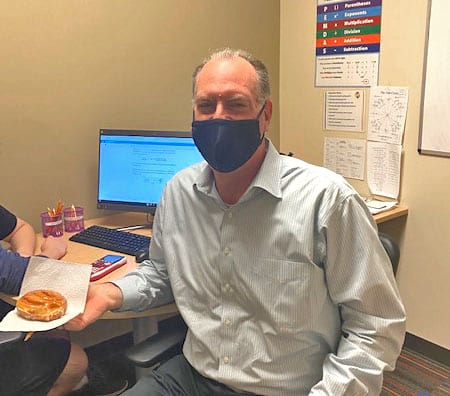 A man wearing a mask is sitting at a desk holding a plate of food.