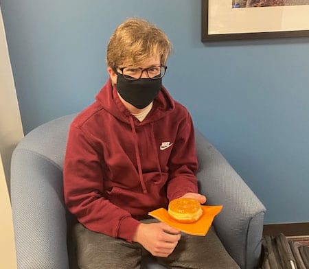 A young man wearing a mask is sitting in a chair holding a donut.