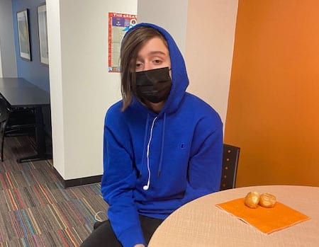 A young man wearing a blue hoodie and a black mask is sitting at a table.