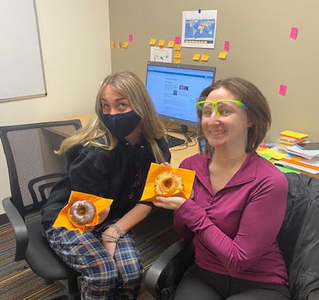 Two women wearing masks are sitting in an office holding donuts.
