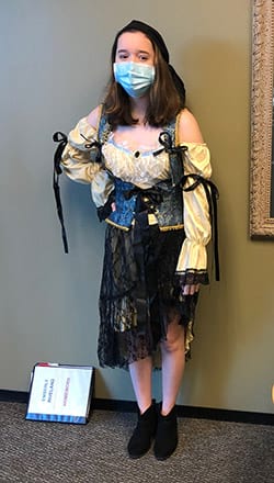 A woman wearing a mask and a pirate costume is standing in front of a wall.