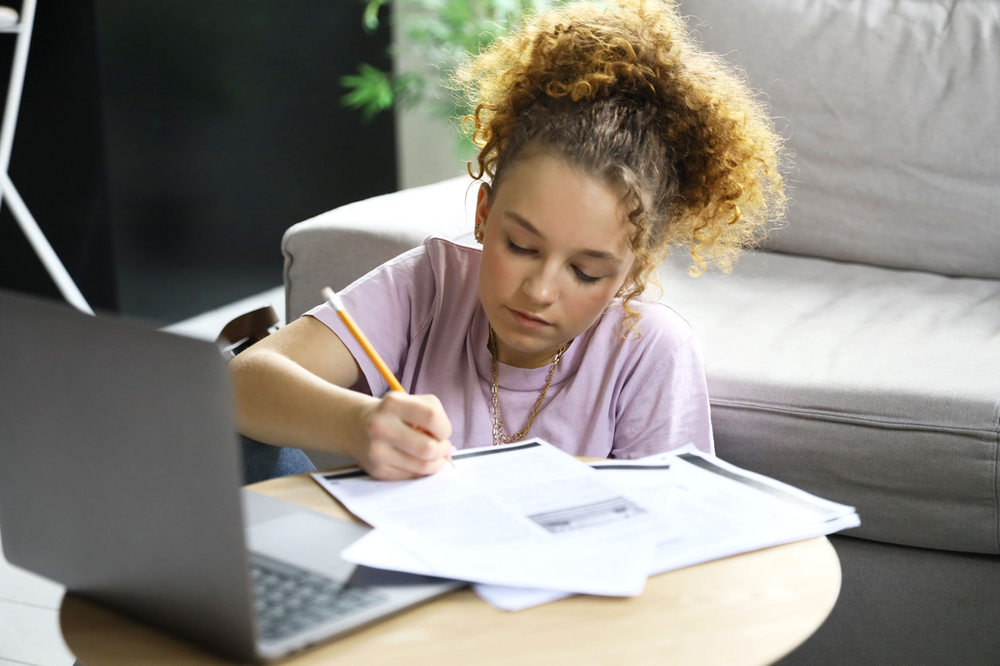 A young girl is sitting at a table with a laptop and writing on a piece of paper.