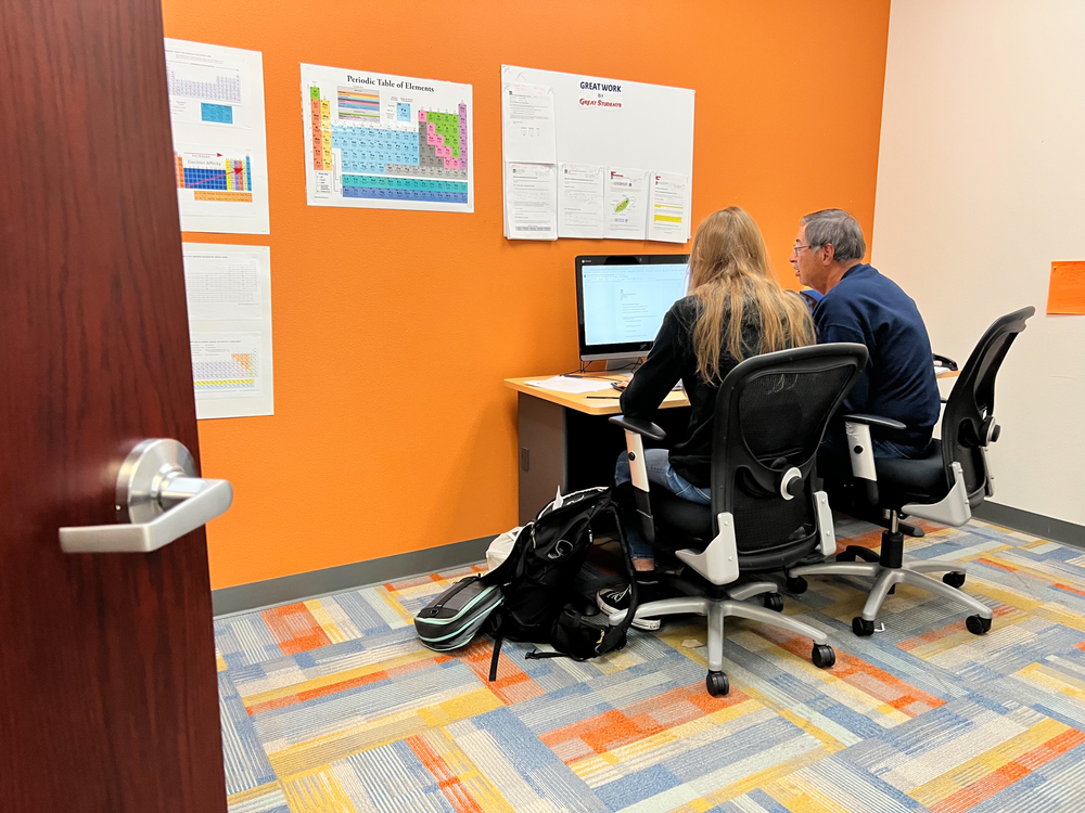 A man and a woman are sitting at a desk in a room with a computer.