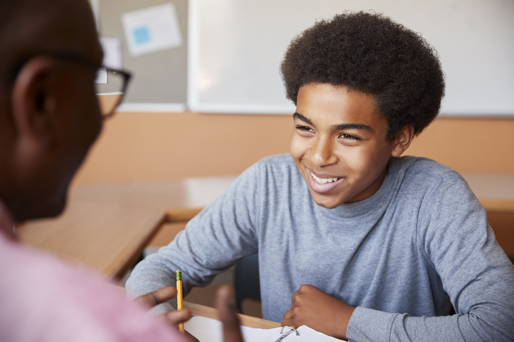 A young boy is sitting at a desk in a classroom talking to a teacher.