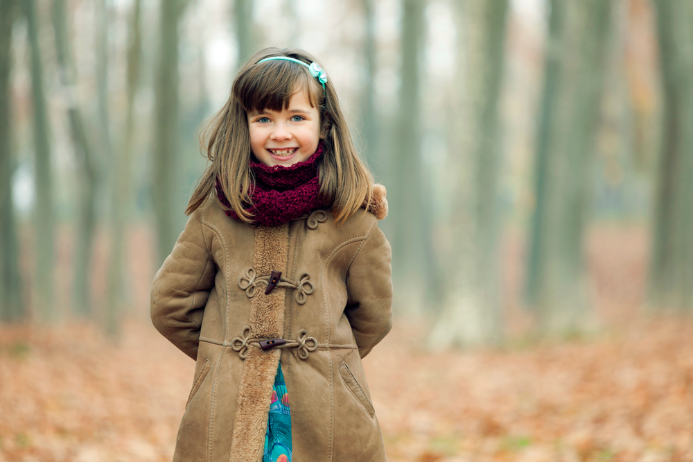 A little girl wearing a coat and scarf is standing in the woods.
