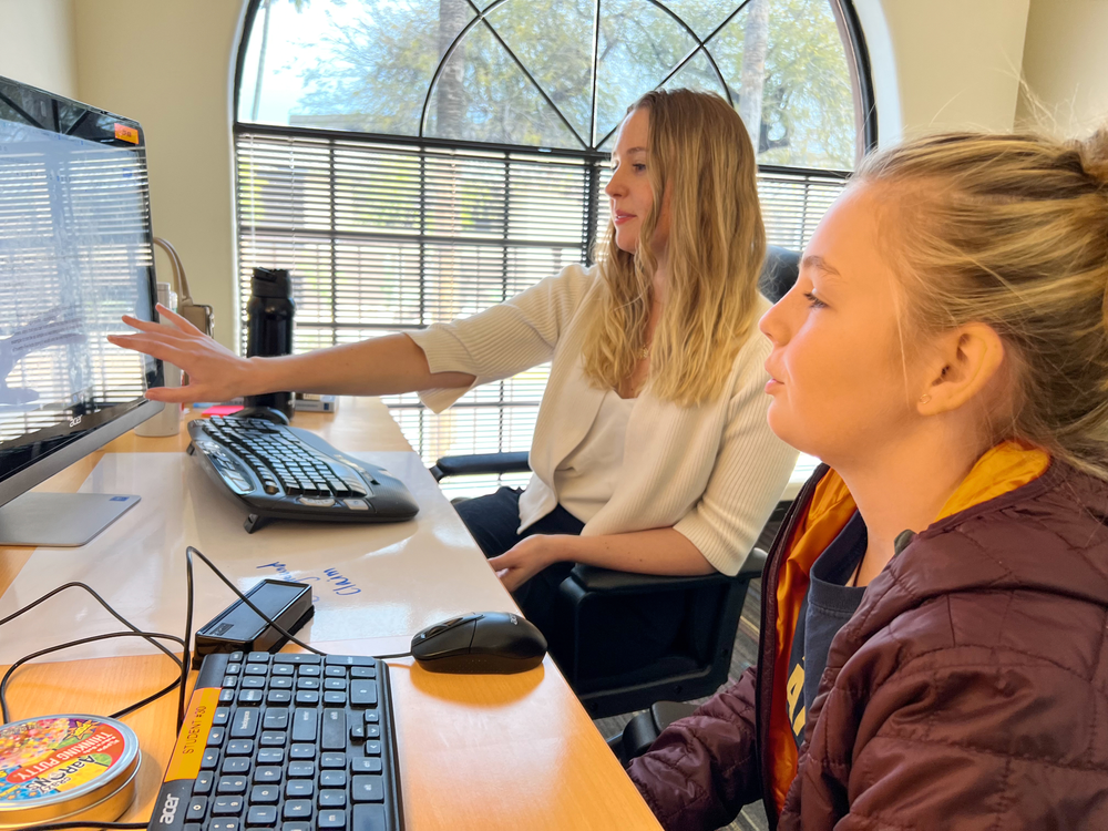 Two women are sitting at a desk looking at a computer screen.