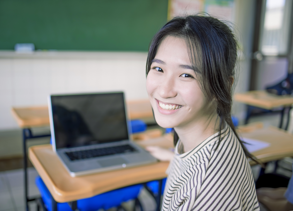A young woman is smiling in a classroom with a laptop on her desk.
