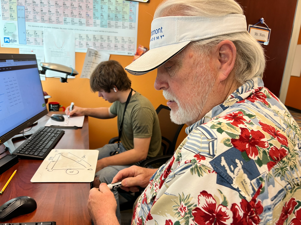A man in a floral shirt is sitting at a desk in front of a computer.