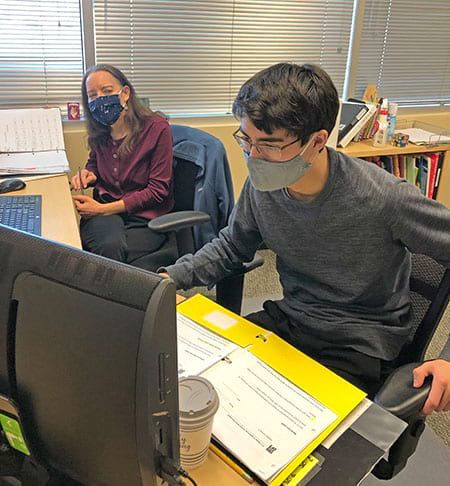A man and a woman wearing masks are sitting at a desk in front of a computer.