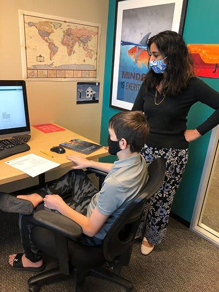 A woman wearing a mask is standing next to a boy sitting at a desk.