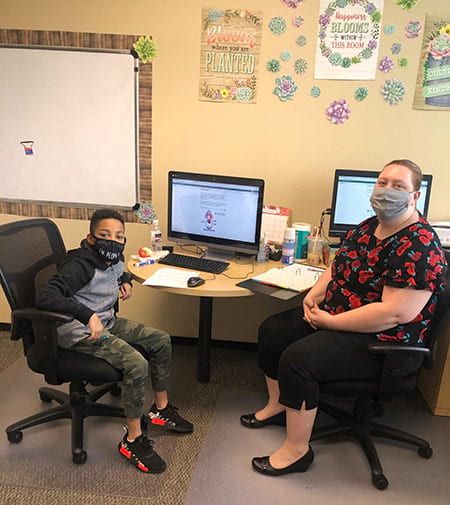 A woman and a boy wearing masks are sitting at a desk with computers.