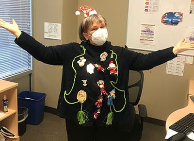 A woman wearing a mask and a christmas sweater is standing in an office with her arms outstretched.