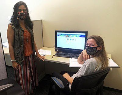 Two women wearing masks are standing next to each other in front of a computer.