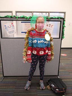 A woman wearing a christmas sweater and a mask is standing in an office cubicle.