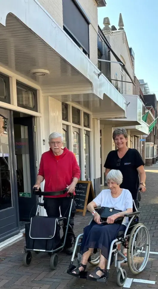 Drie mensen staan buiten een gebouw; één gebruikt een rollator, een ander zit in een rolstoel en de derde duwt de rolstoel.