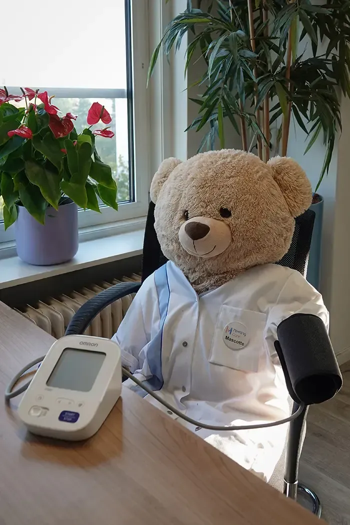 Een teddybeer in een witte mantel zit aan een tafel met een bloeddrukmeter bij een raam en potplanten.