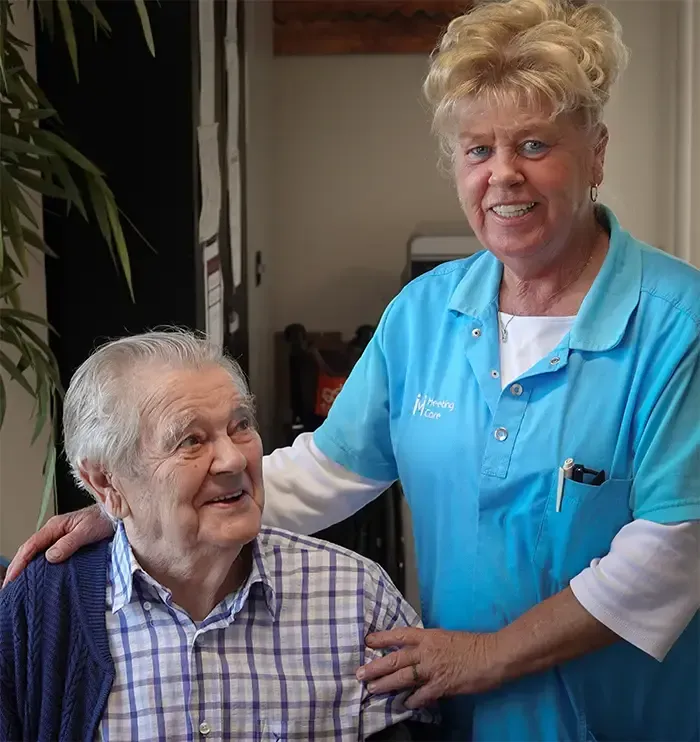 Verzorgster in een blauwe polo glimlacht naast een zittende man in een geruit overhemd in een huiselijke omgeving.