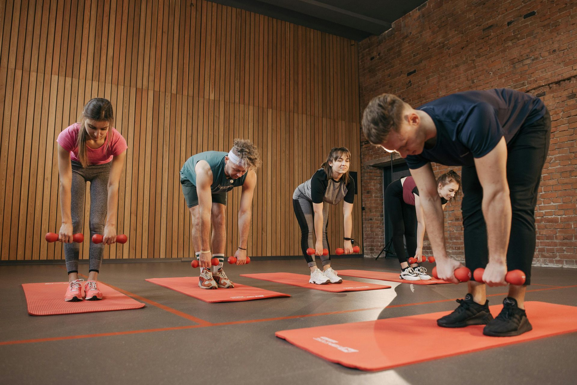 People exercising in a studio, bending forward with dumbbells. Wooden wall and red mats are visible.