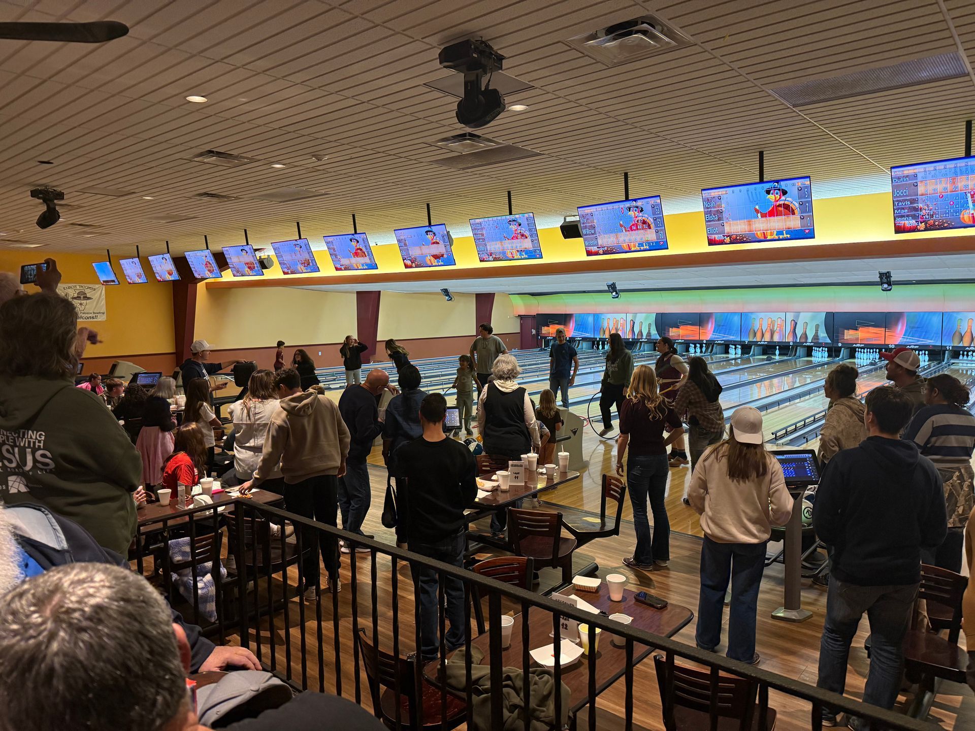 Busy bowling alley with people seated and standing by lanes under colorful overhead score screens.