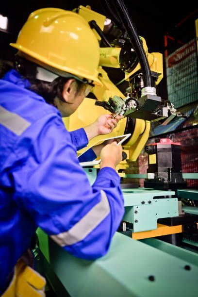 Factory worker in blue overalls and yellow helmet examines machinery with a tablet.