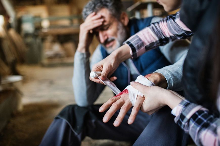 Man bandages another person's hand after a cut, with an injured man sitting nearby in the background.
