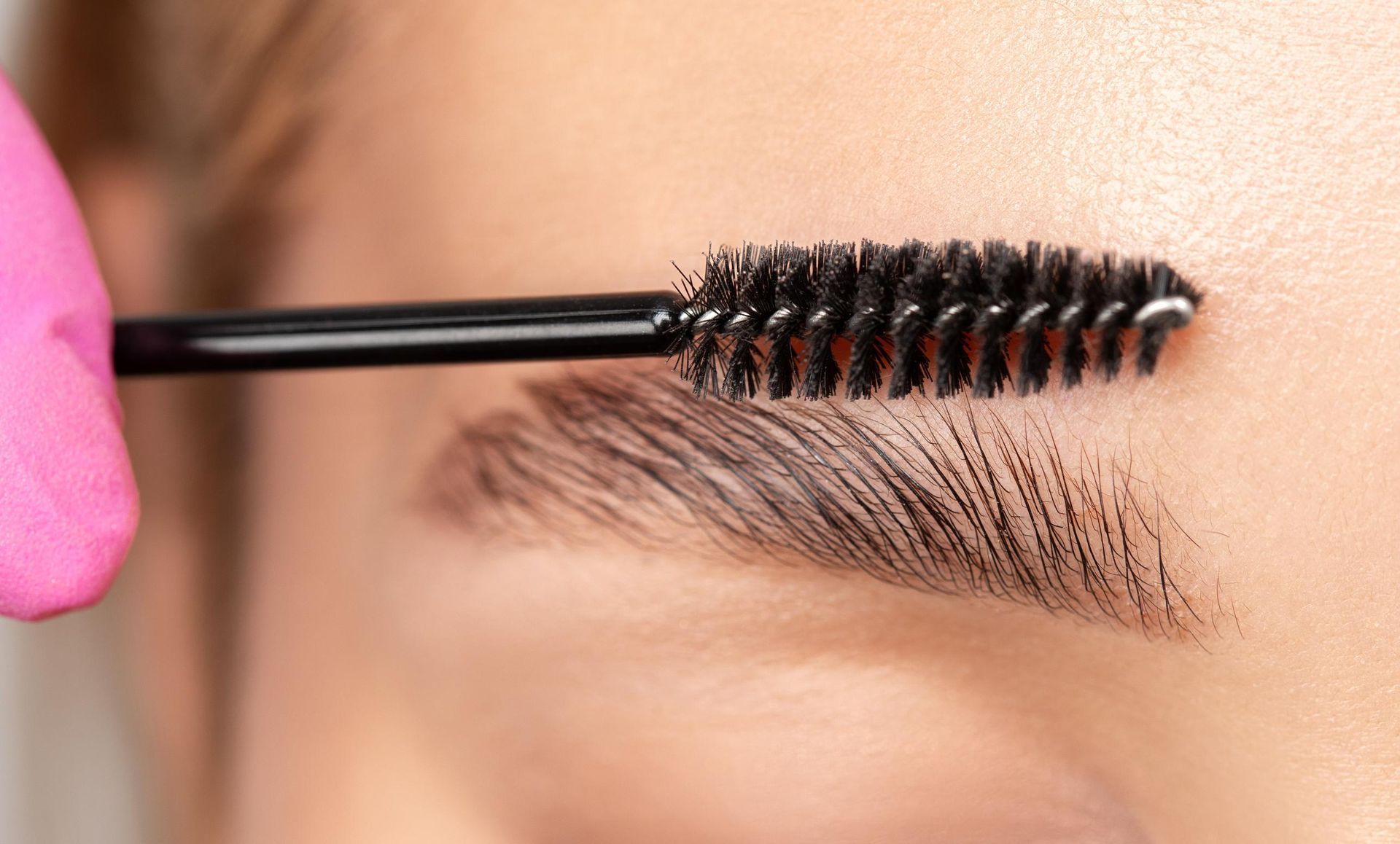 A woman is applying mascara to her eyebrows with a pink brush.