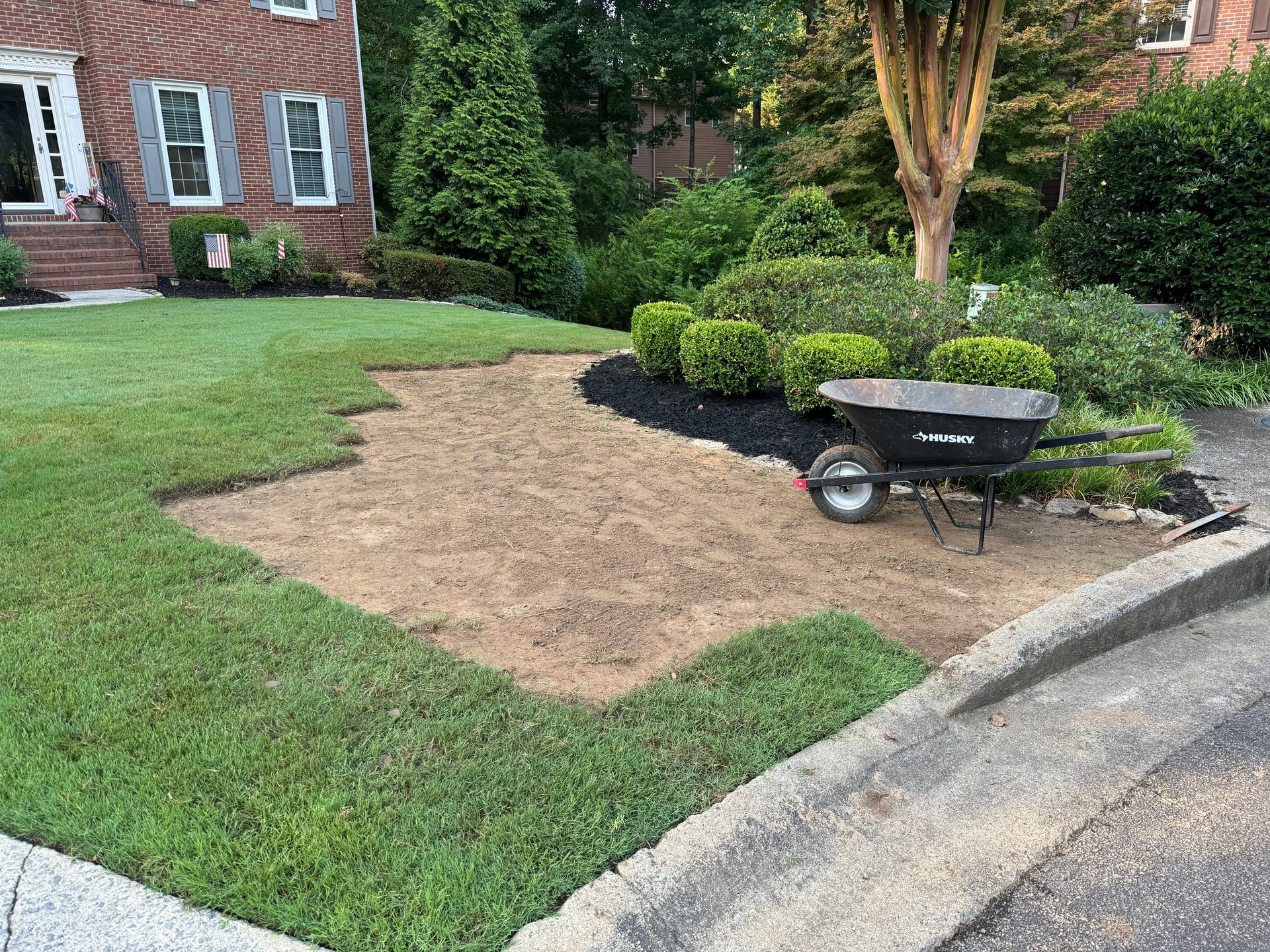 Mulch-covered lawn patch beside a brick house, with a wheelbarrow near the curb and shrubs in the background