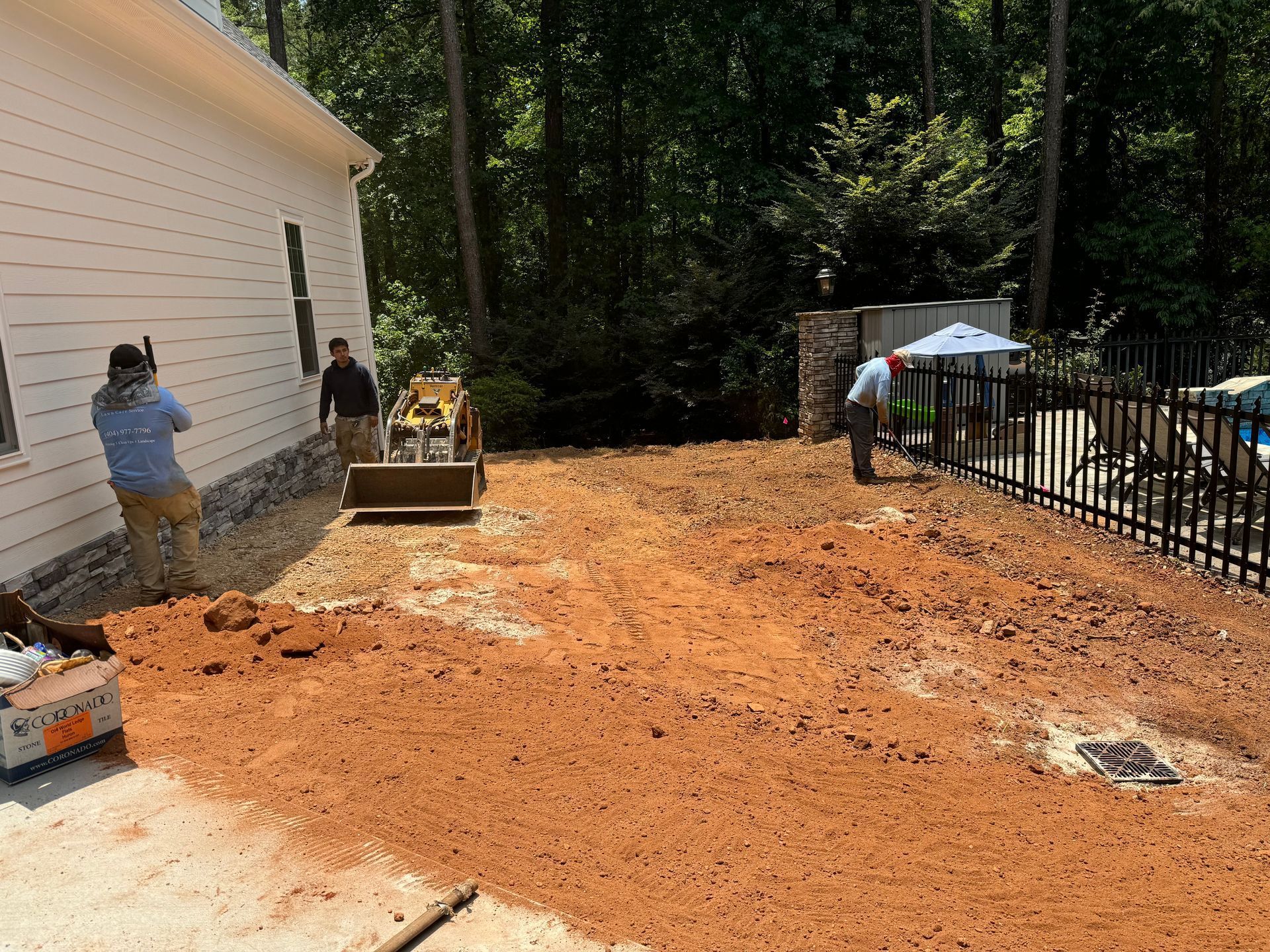 Workers leveling reddish dirt beside a white house and black fence in a wooded yard