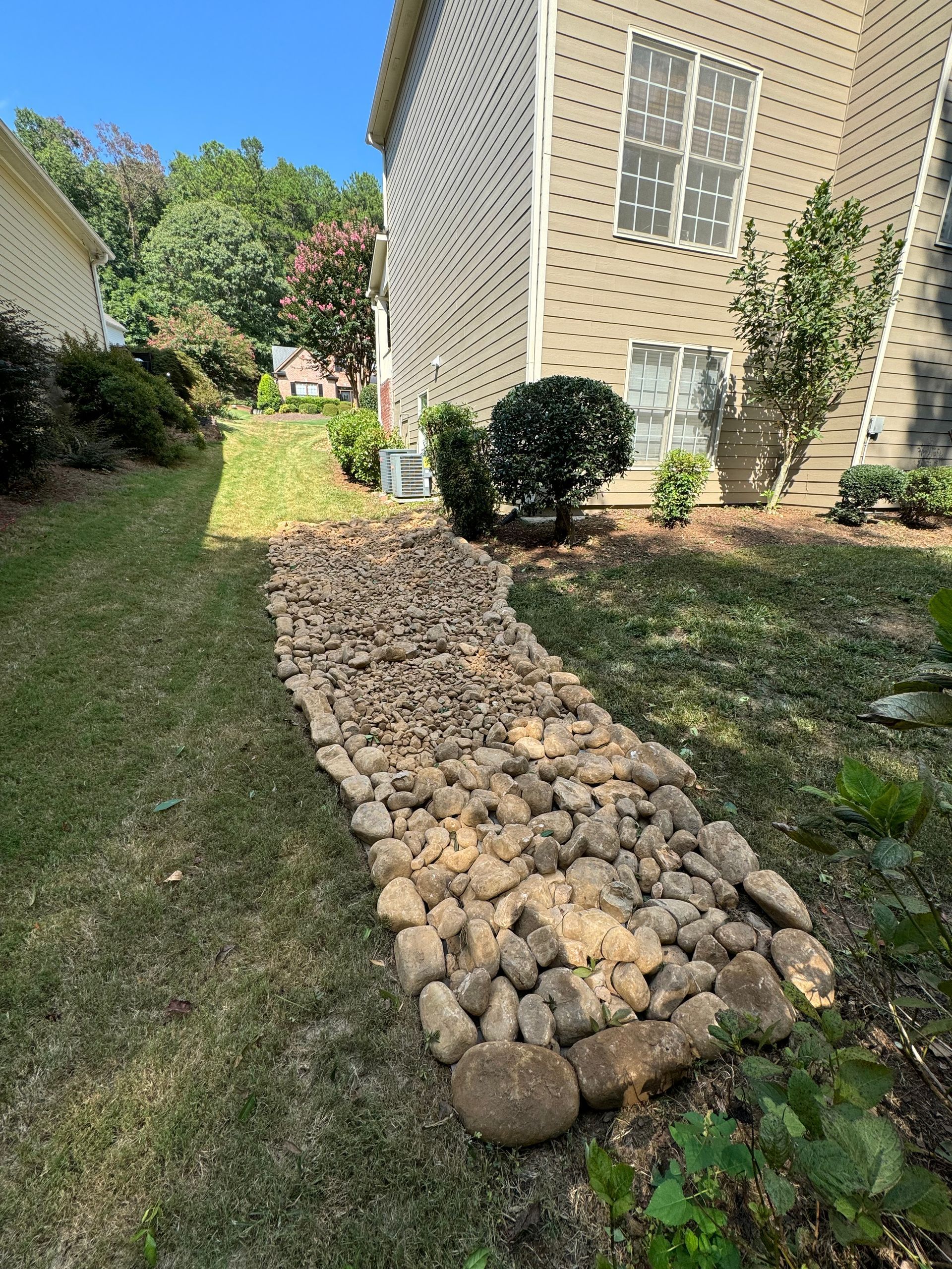 Rock-lined garden border beside a house with grass and shrubs under a sunny sky