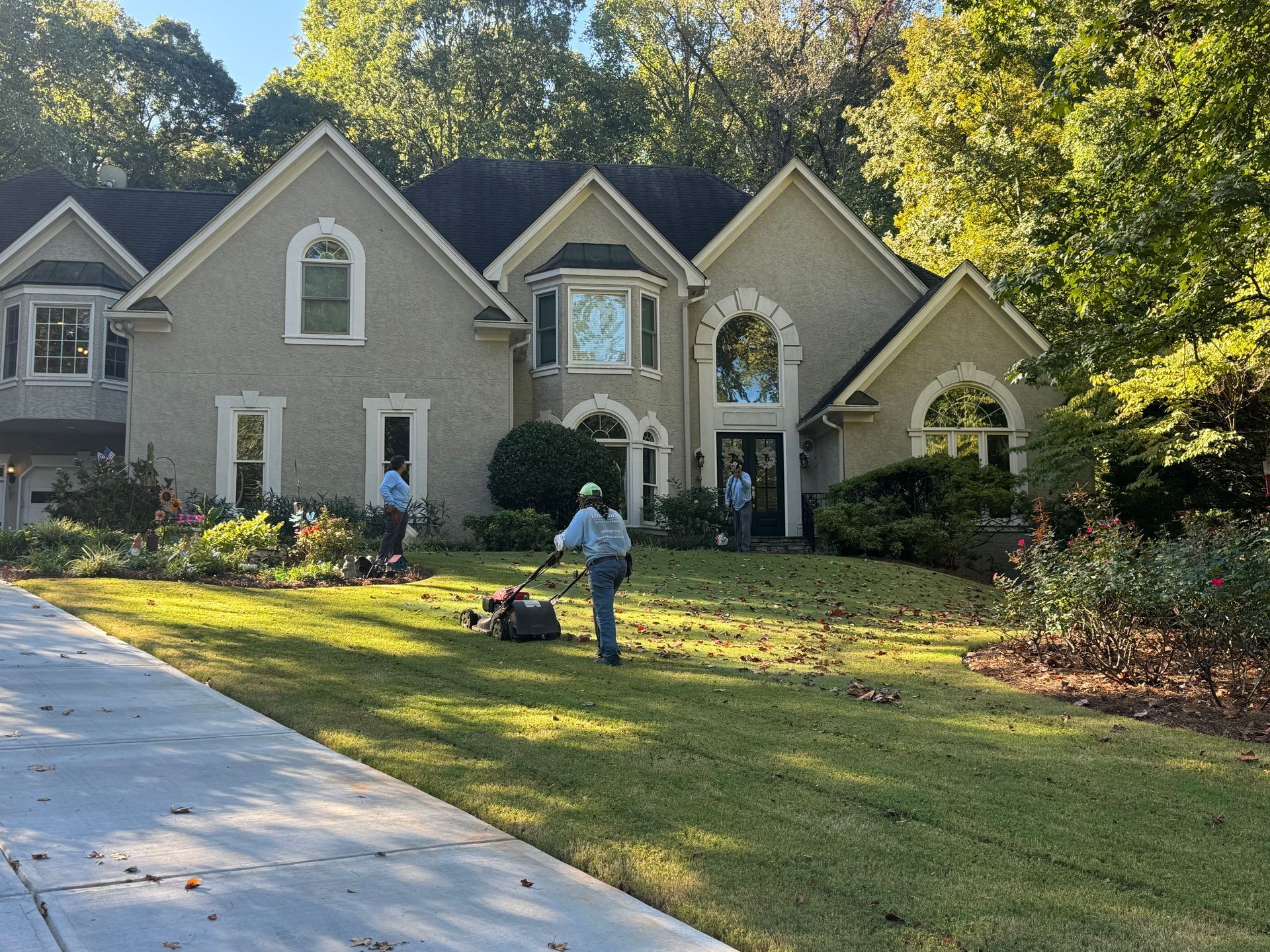 Person mowing lawn in front of a gray suburban house with arched windows and trees.