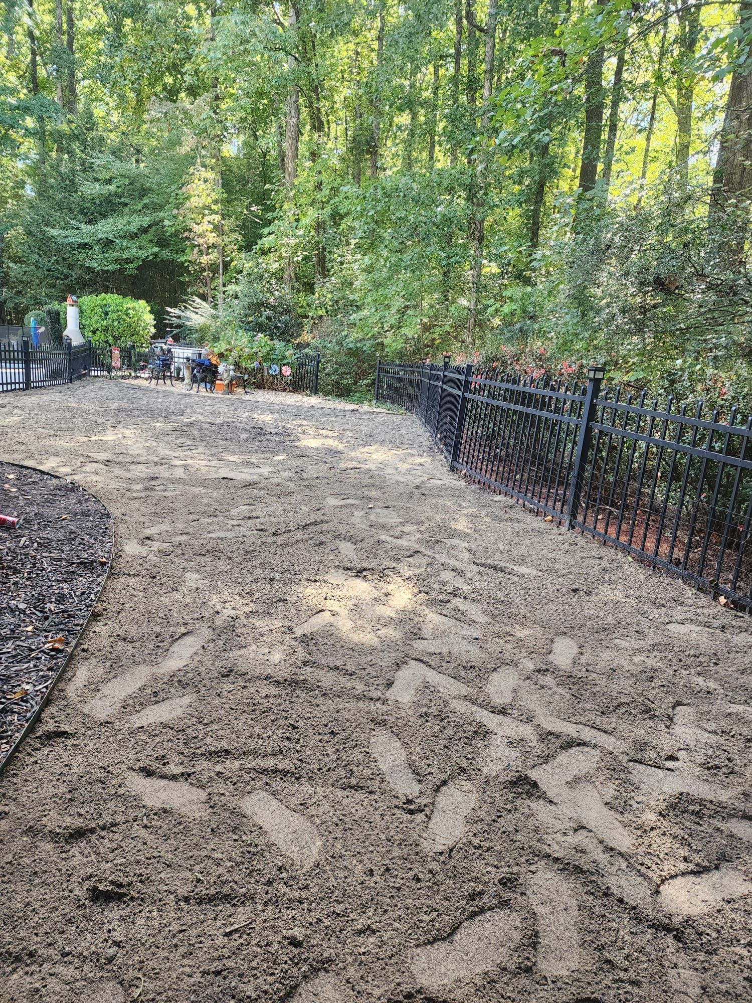 Sandy pathway beside a black fence, leading through a wooded garden area in daylight.