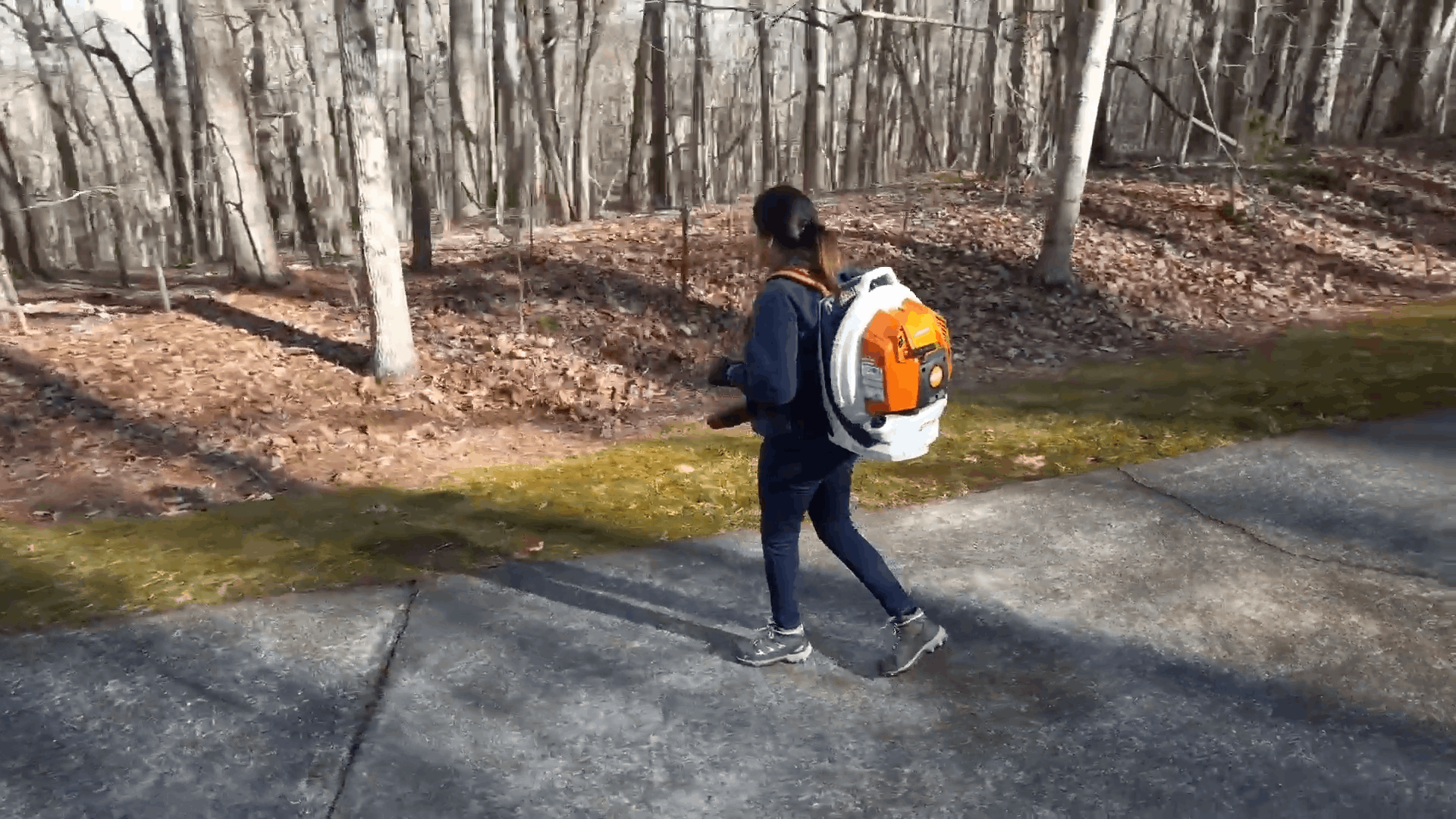 Person with orange backpack walking on a paved path beside a wooded area in winter
