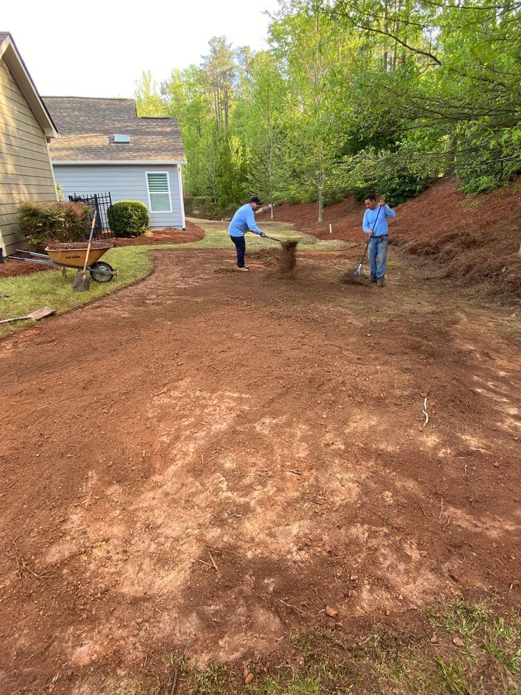 Two workers leveling a dirt yard beside a house, with a wheelbarrow and trees in the background.