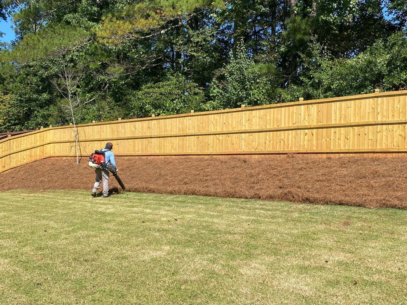 Two people mowing a grassy lawn beside a wooden fence and brick retaining wall