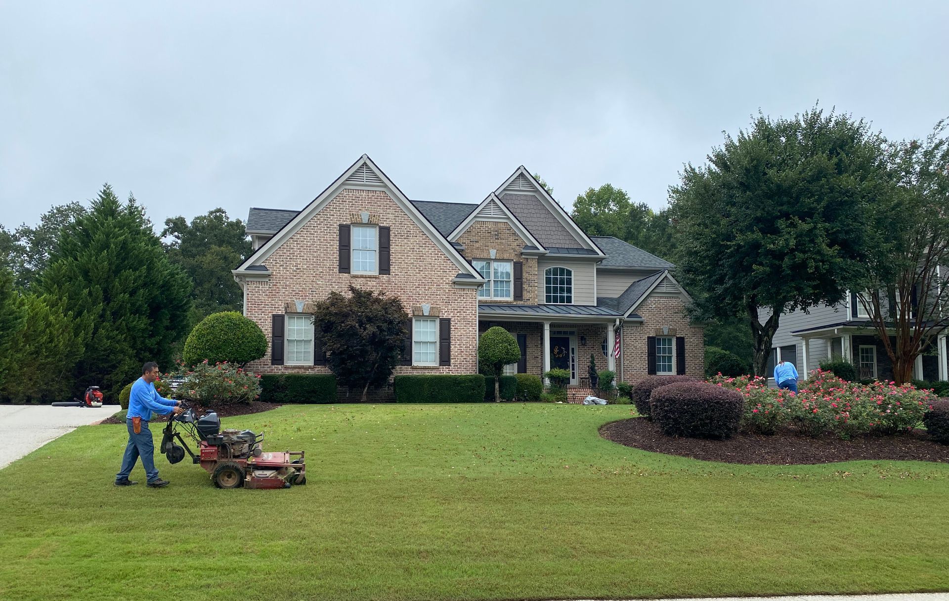 Person mowing a large front lawn in front of a brick suburban house on an overcast day