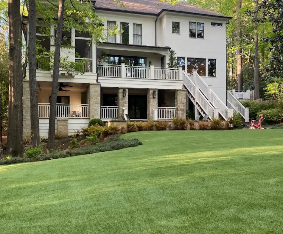 Two-story white house with a large deck and staircase, set behind a green lawn and trees.