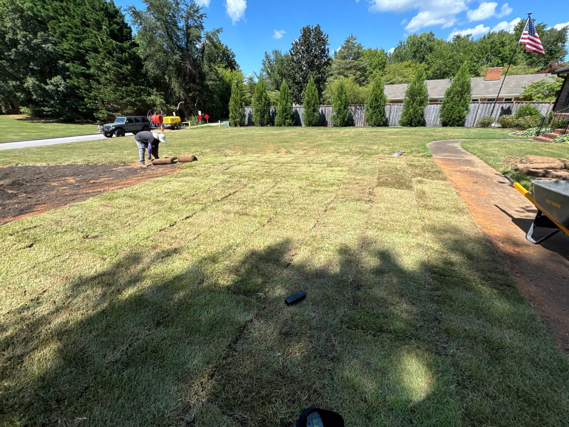 Person mowing a large grassy yard on a sunny day with trees and a house in the background