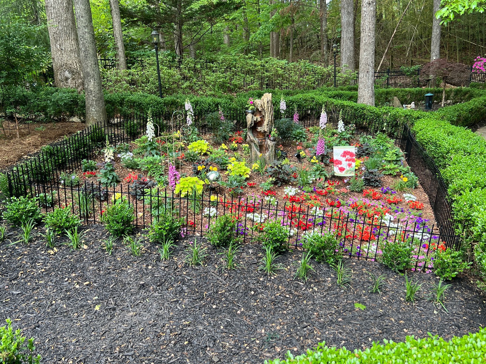 Colorful flower garden bed bordered by black fencing in a wooded park.