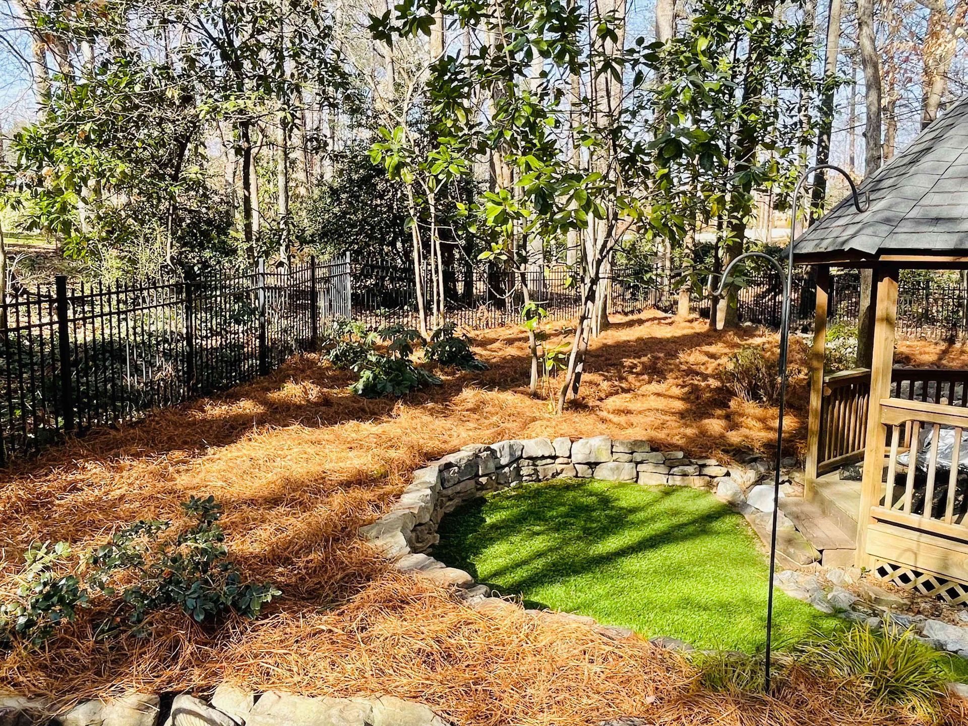 Backyard with pine straw, a curved stone flower bed, green grass, and a wooden gazebo by trees