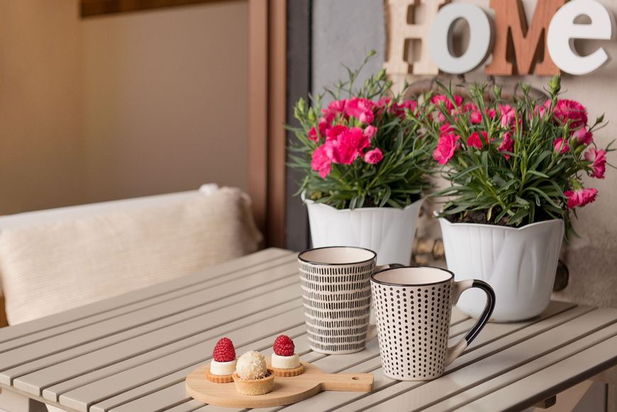 Dettaglio ravvicinato di un tavolino sul balcone con due tazze decorate, piccoli dolcetti e vasi di fiori rosa sullo sfondo.