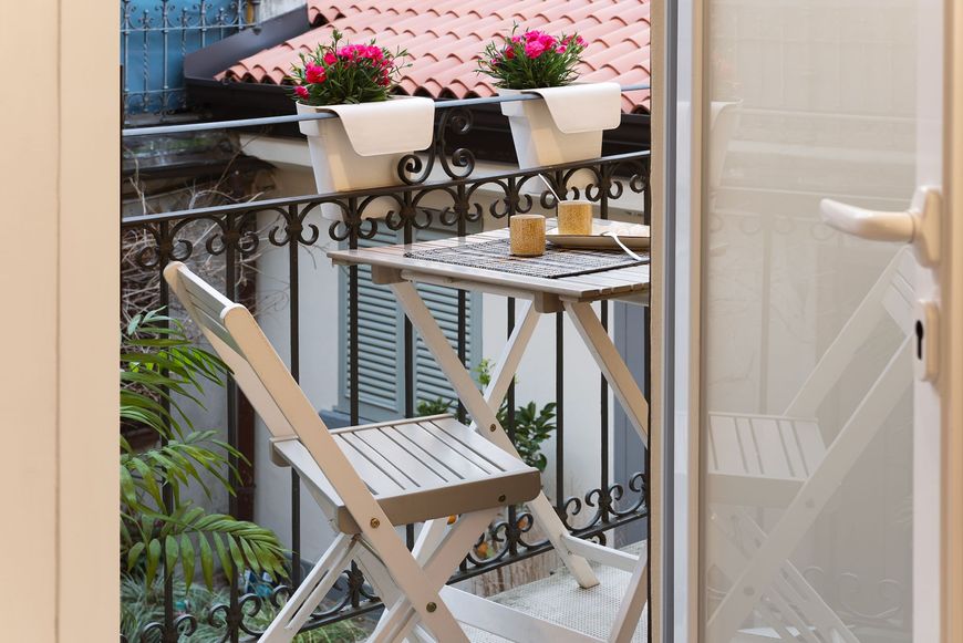 Piccolo balcone esterno attrezzato con un tavolino e due sedie pieghevoli in legno, con vasi di fiori appesi alla ringhiera.