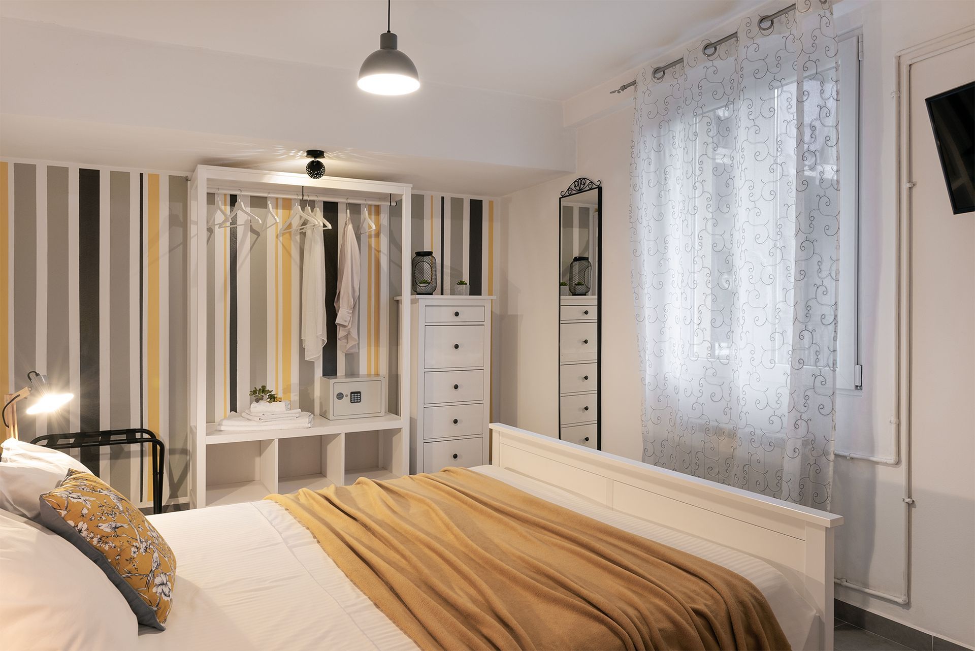 Bright bedroom with a double bed, gold bed runner, striped wallaper, open wardrobe, and a window with a white curtain.