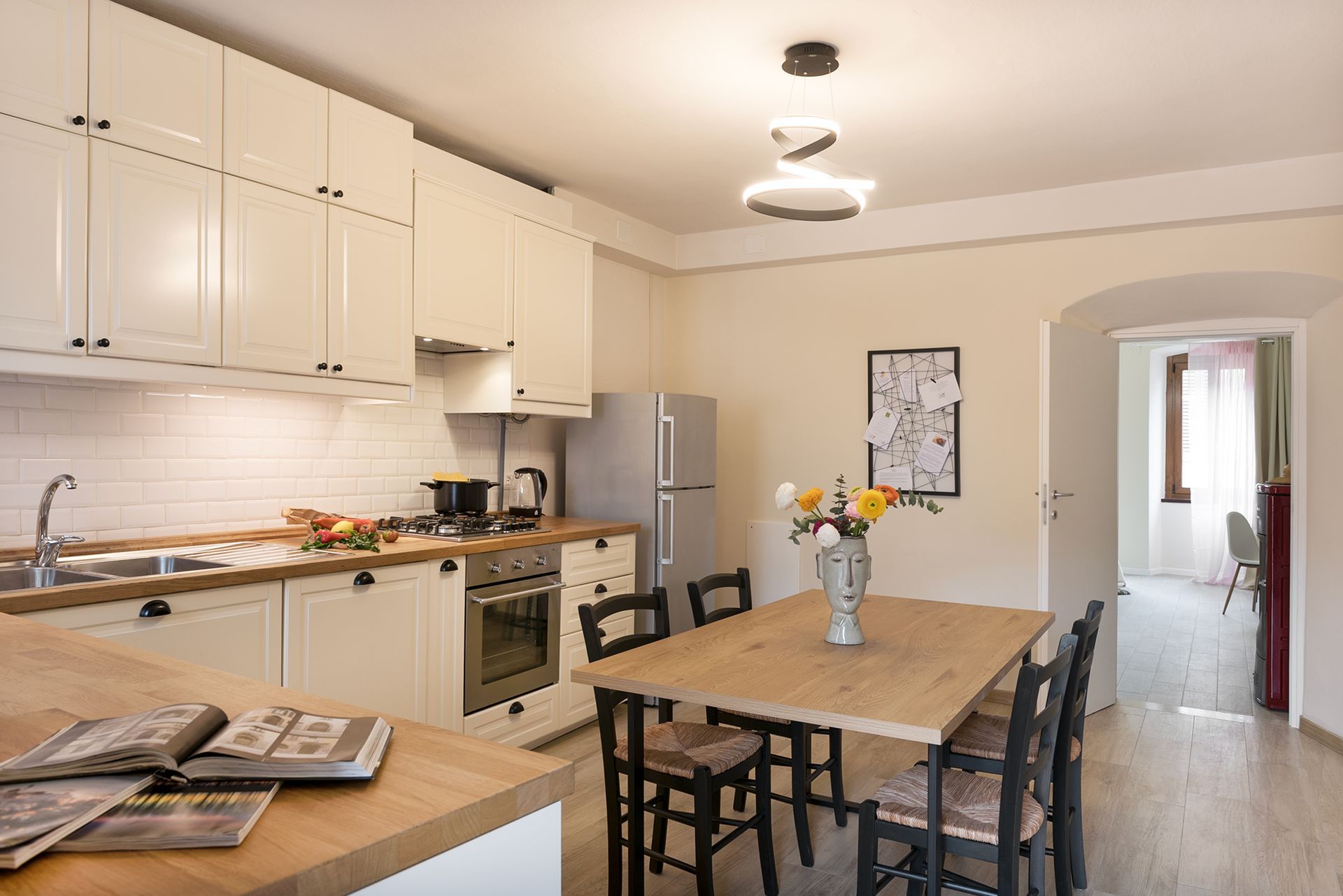 Large white kitchen showing the wooden table with a vase of yellow flowers and a view towards the entrance hallway.