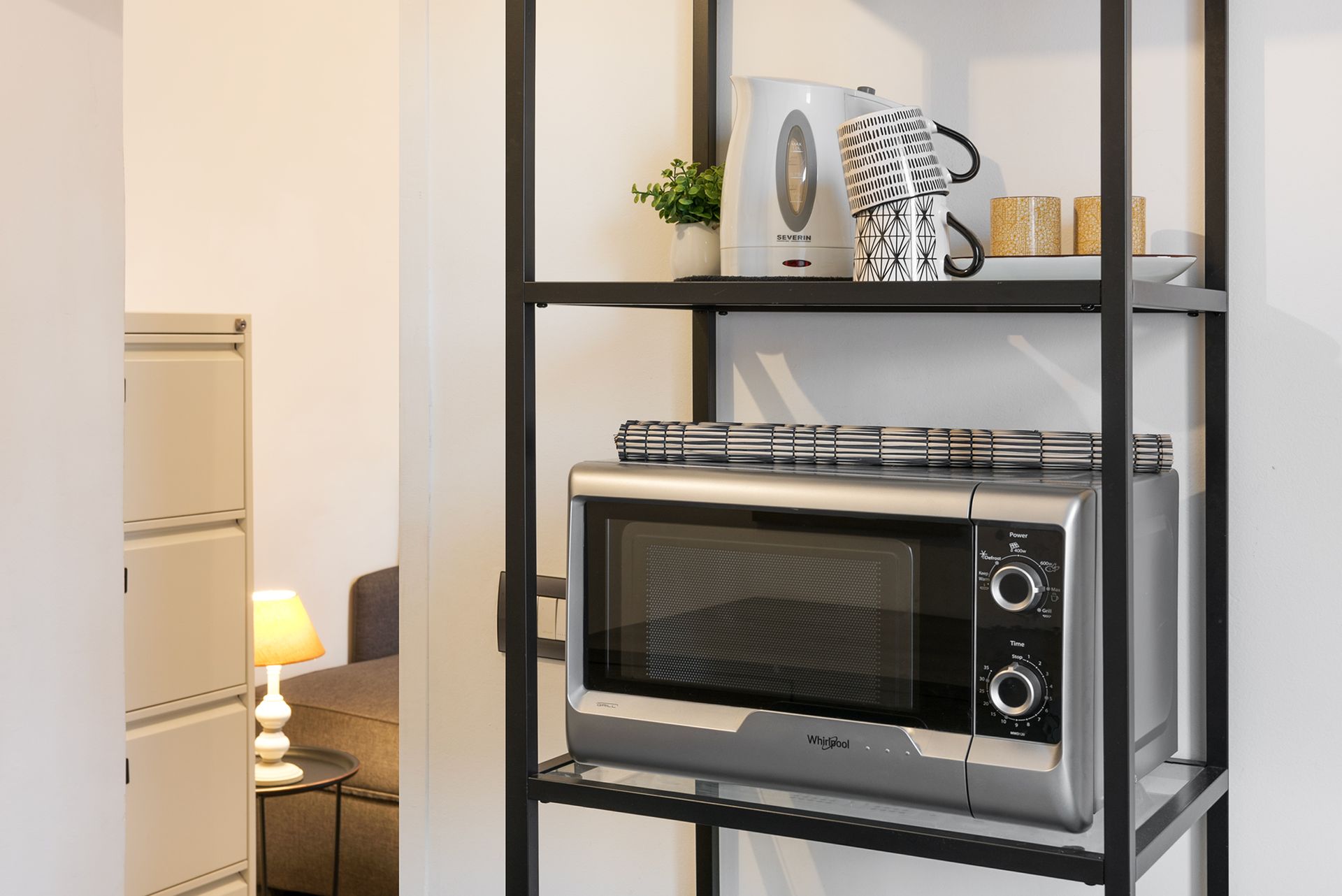 Detail of a black metal shelving unit in the kitchen containing a microwave oven and coffee mugs.