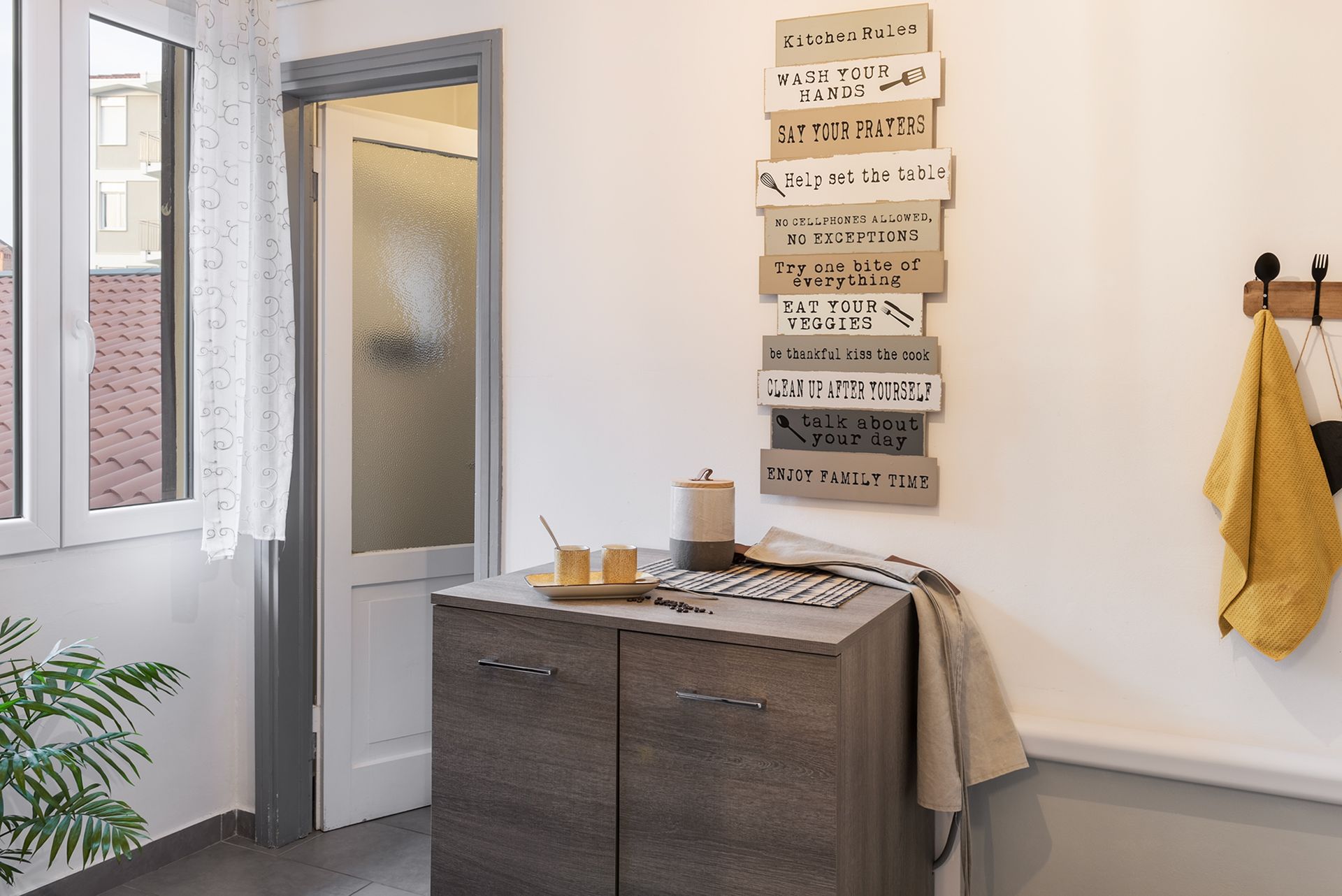 View of a room with a window, a dark wood chest of drawers, wall decorations, and a yellow tea towel.