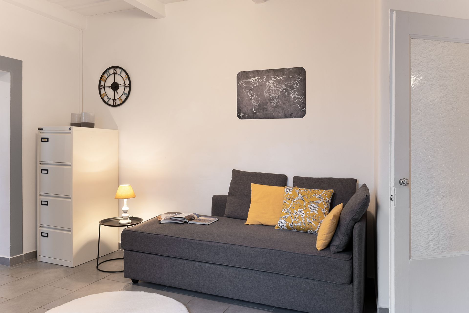 Living room with a grey sofa bed, yellow pillows, a tall white chest of drawers, a lit table lamp, and a wall clock.